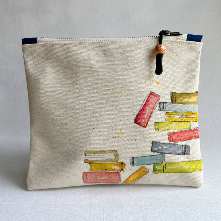 An off-white canvas zippered pouch featuring a hand-painted illustration of colorful books on the front, with a black and orange tassel zipper pull, set against a neutral light gray background.