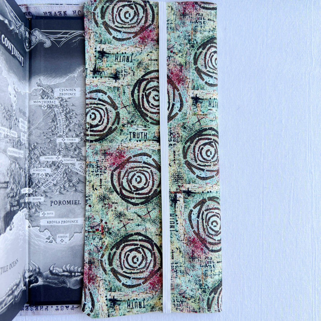a book covered in a vibrant blue green fabric reusable  adjustable book cover.  Fabric has impressionist rose / concentric deep brown circles and the word ' TRUTH scattered on the pattern.