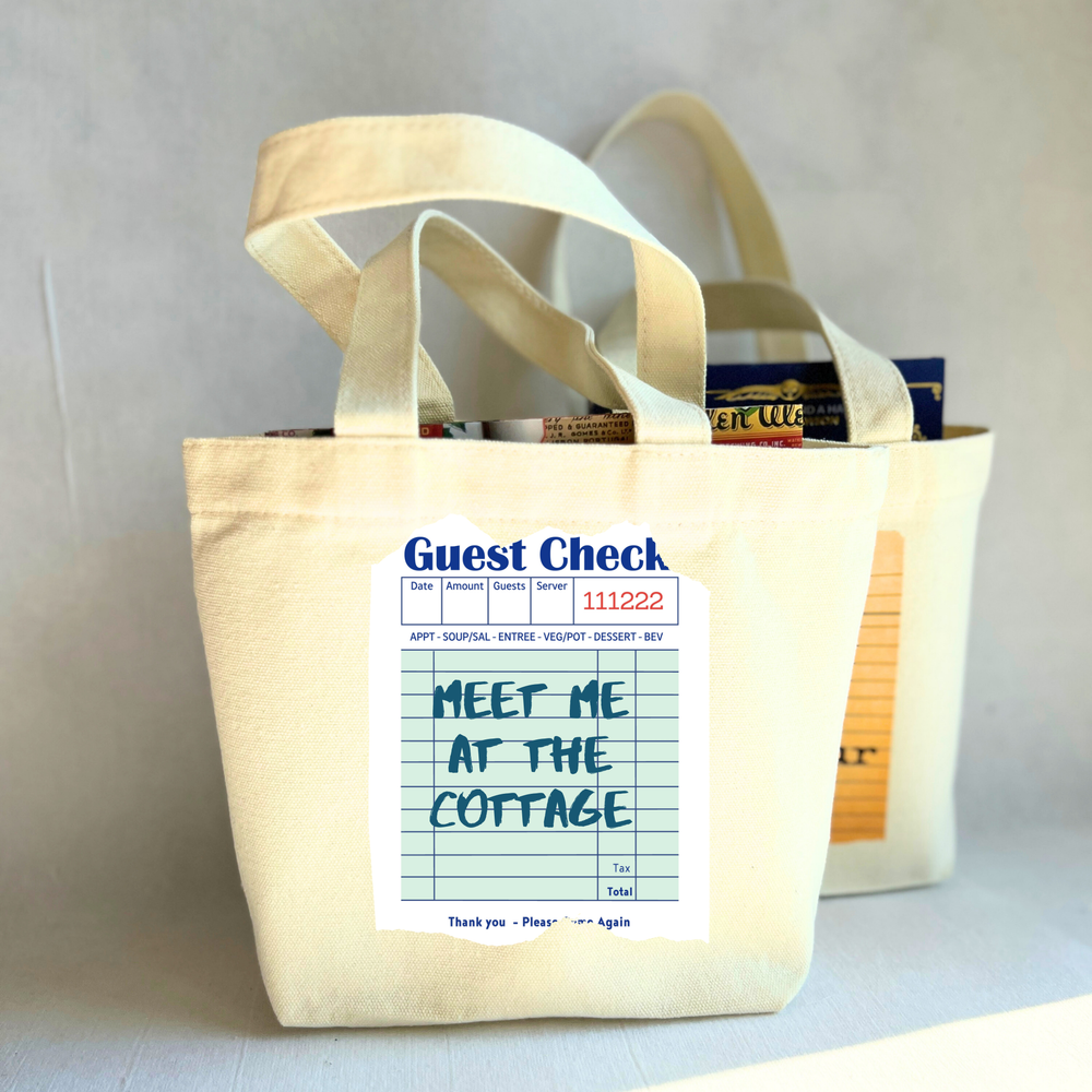 Beige small tote bag with a printed guest check label on a light green background with the words, 'Meet me at the cottage.'