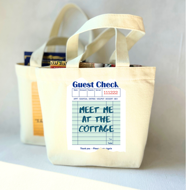 Beige small tote bag with a printed guest check label on a light green background with the words, 'Meet me at the cottage.'