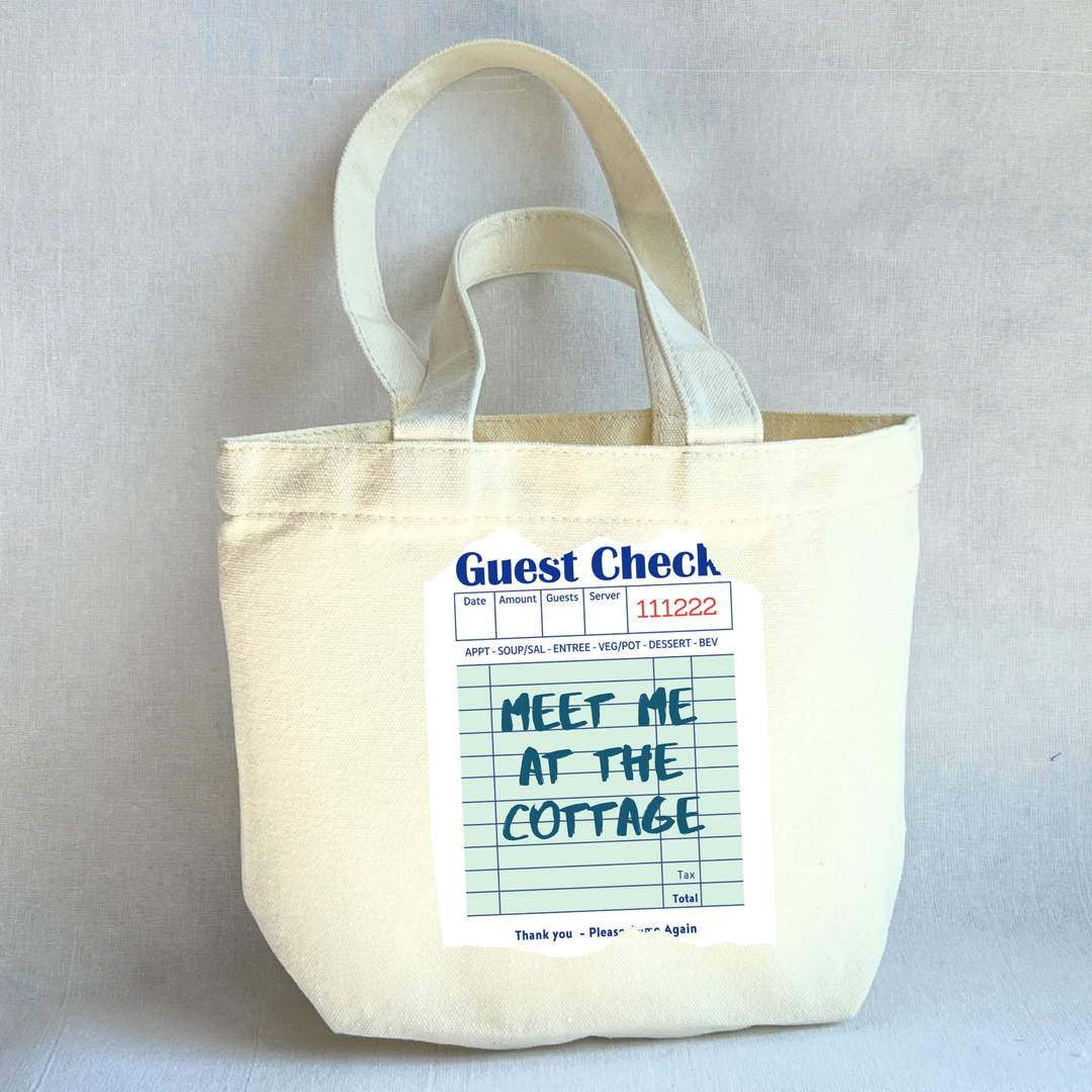 Beige small tote bag with a printed guest check label on a light green background with the words, 'Meet me at the cottage.'