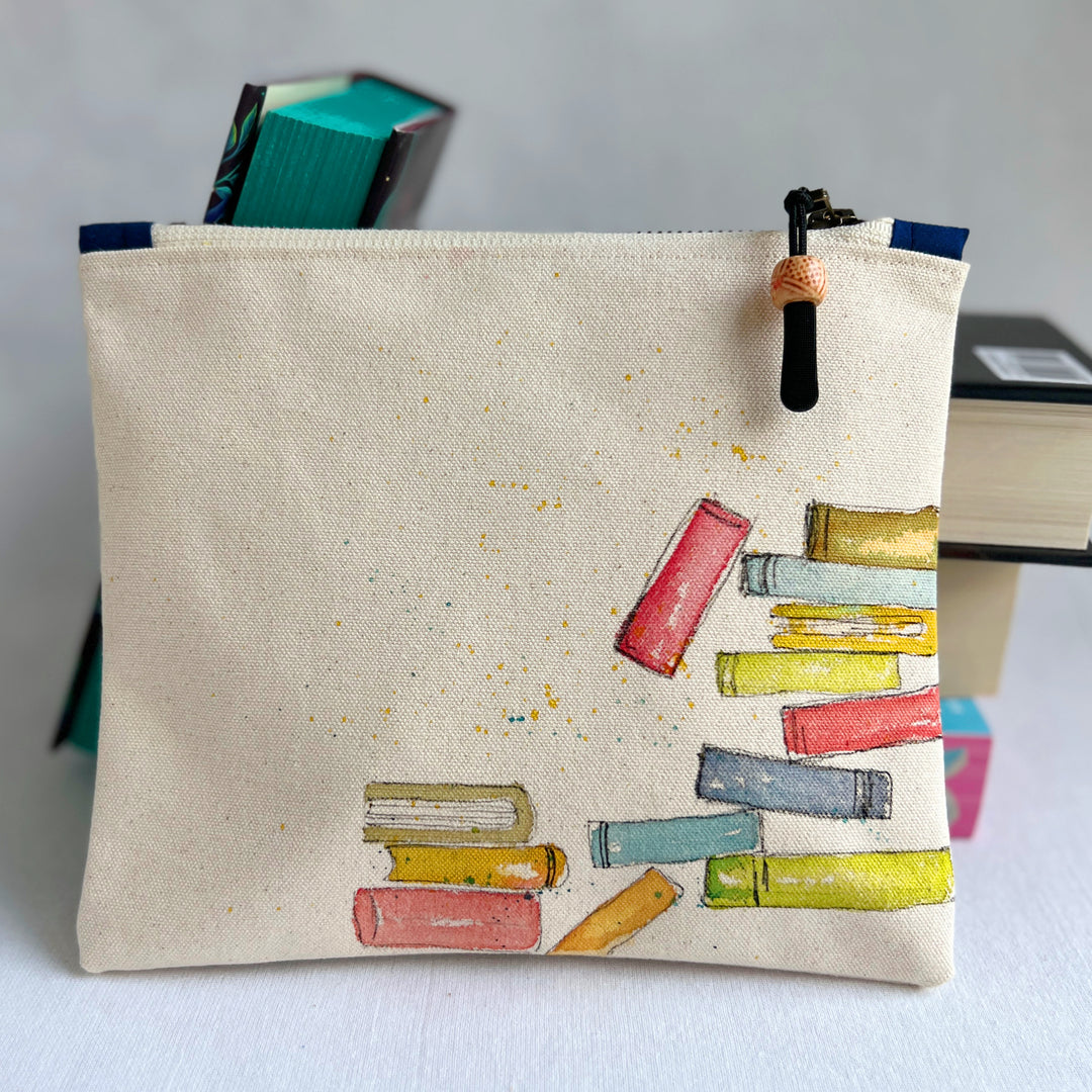 An off-white canvas zippered pouch featuring a hand-painted illustration of colorful books on the front, with a black and orange tassel zipper pull, set against a neutral light gray background.