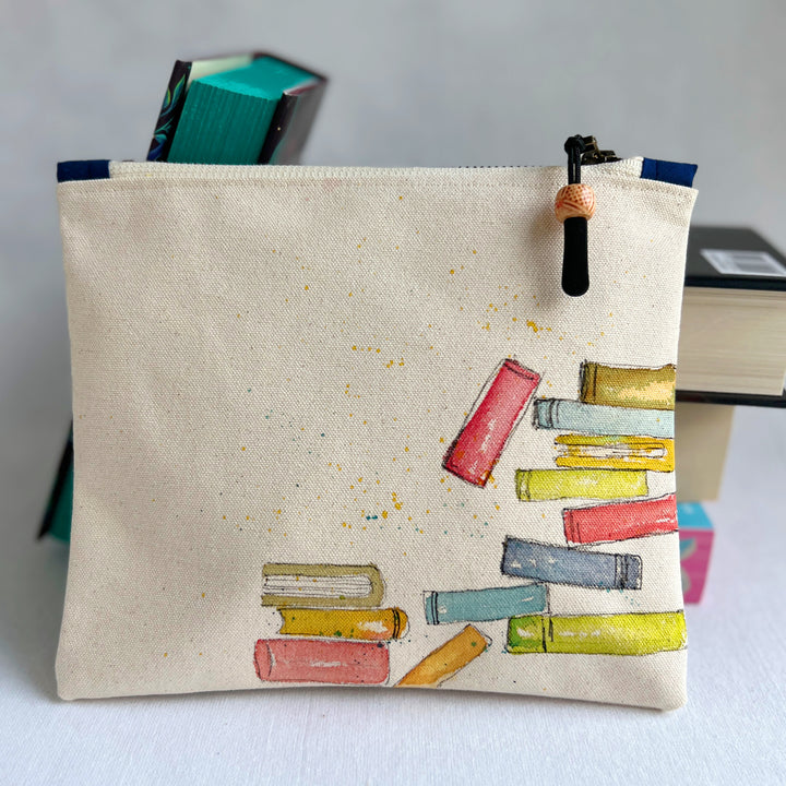 An off-white canvas zippered pouch featuring a hand-painted illustration of colorful books on the front, with a black and orange tassel zipper pull, set against a neutral light gray background.