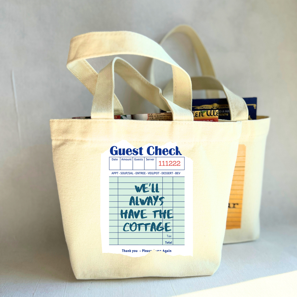 Beige small tote bag with a printed guest check label on a light green background with the words, 'We'll always have the cottage.'