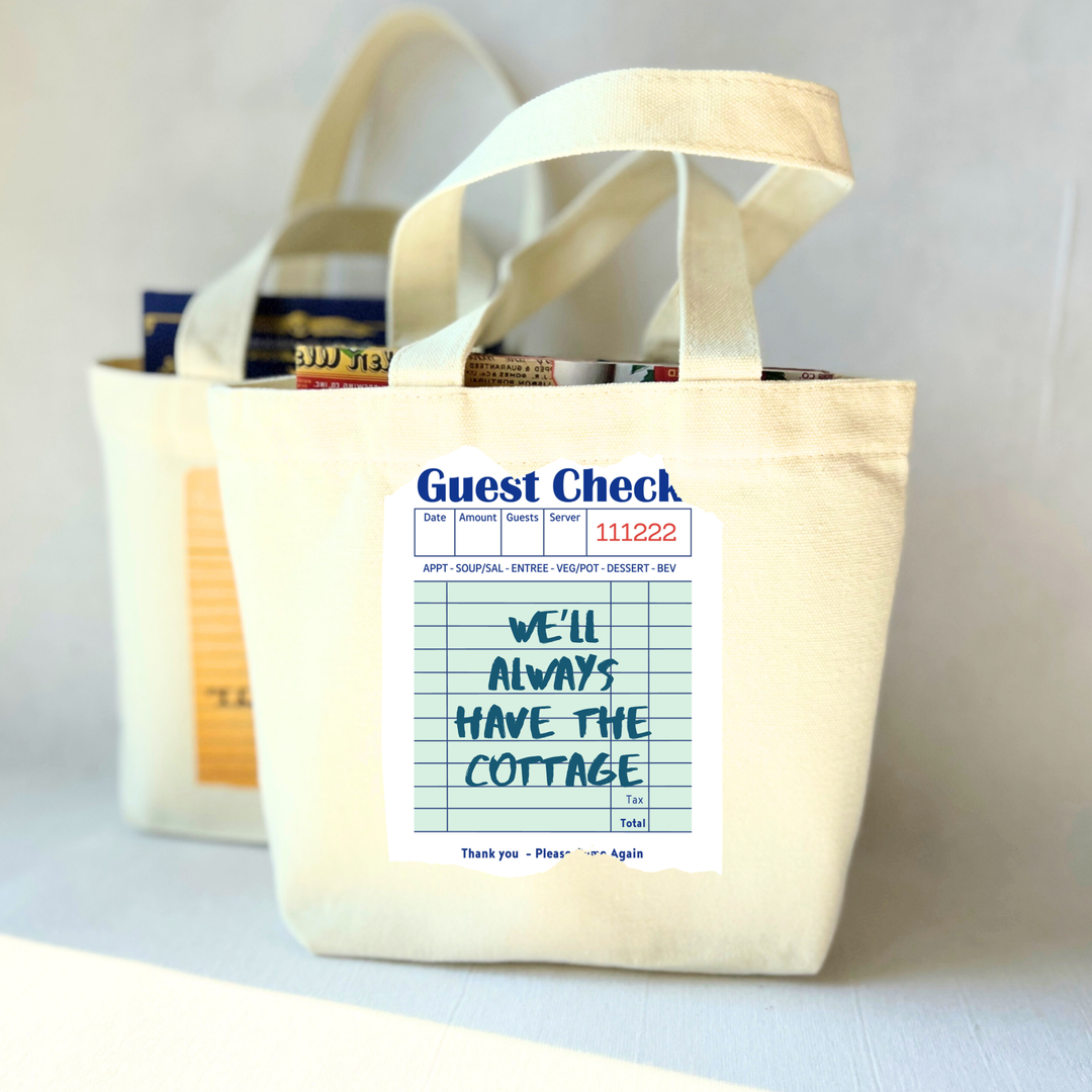 Beige small tote bag with a printed guest check label on a light green background with the words, 'We'll always have the cottage.'