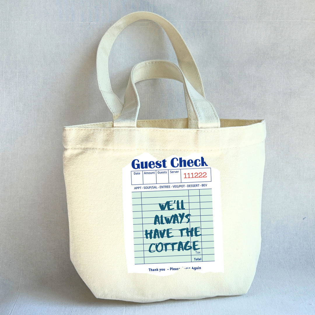 Beige small tote bag with a printed guest check label on a light green background with the words, 'We'll always have the cottage.'