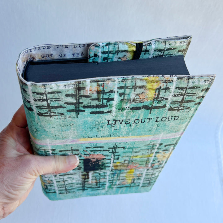 a hand holding a book covered in a vibrant blue green fabric reusable book cover with the words, 'live out loud' printed on the top.