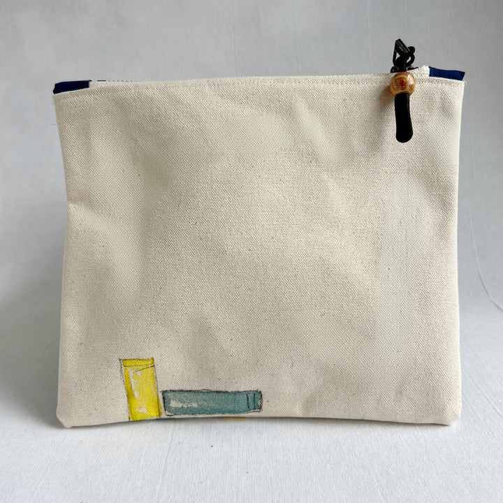 The back side of a square canvas pouch in natural cream canvas with a watercolor-style illustration of colorful book spines along the bottom. The pouch has a top zipper with a black cord pull and a wooden bead accent.