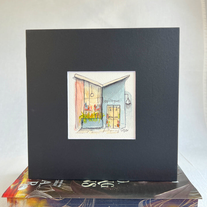 A matted watercolor painting of a cozy midcentury modern bookshop storefront with the store name  'EPILOGUE' above a window filled with books, a glass door, and a hanging lamp, shown on a white mat and white frame.