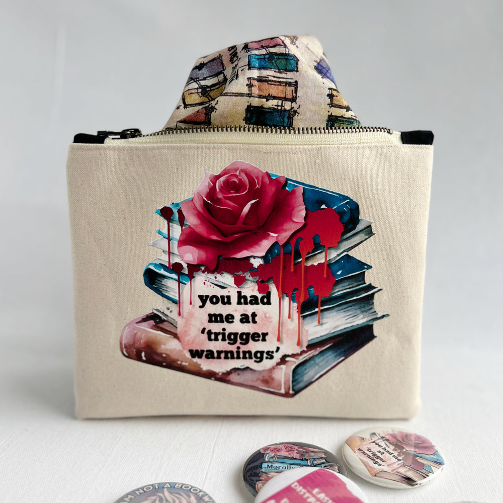 square zippered bag made of canvas, with a design depicting a stack of books, a rose and blood drips, with the words, "you had me at trigger warnings."