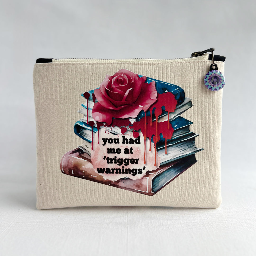 square zippered bag made of canvas, with a design depicting a stack of books, a rose and blood drips, with the words, "you had me at trigger warnings."