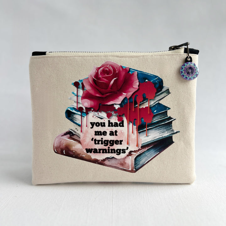square zippered bag made of canvas, with a design depicting a stack of books, a rose and blood drips, with the words, "you had me at trigger warnings."