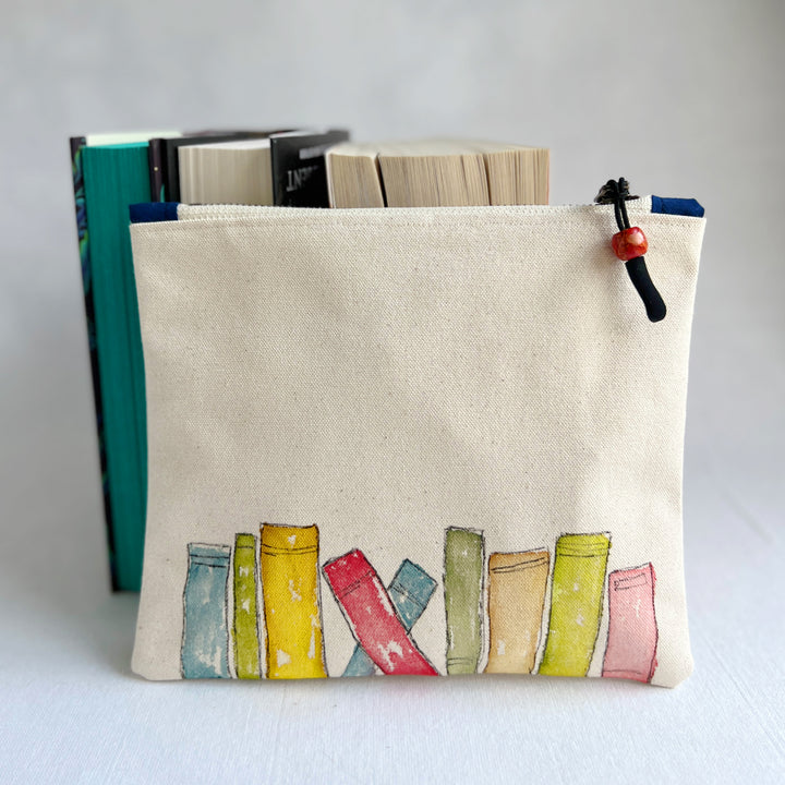 A canvas zippered pouch with a handpainted watercolor of colorful book spines along the bottom, shown standing upright with books behind it against a light gray background.
