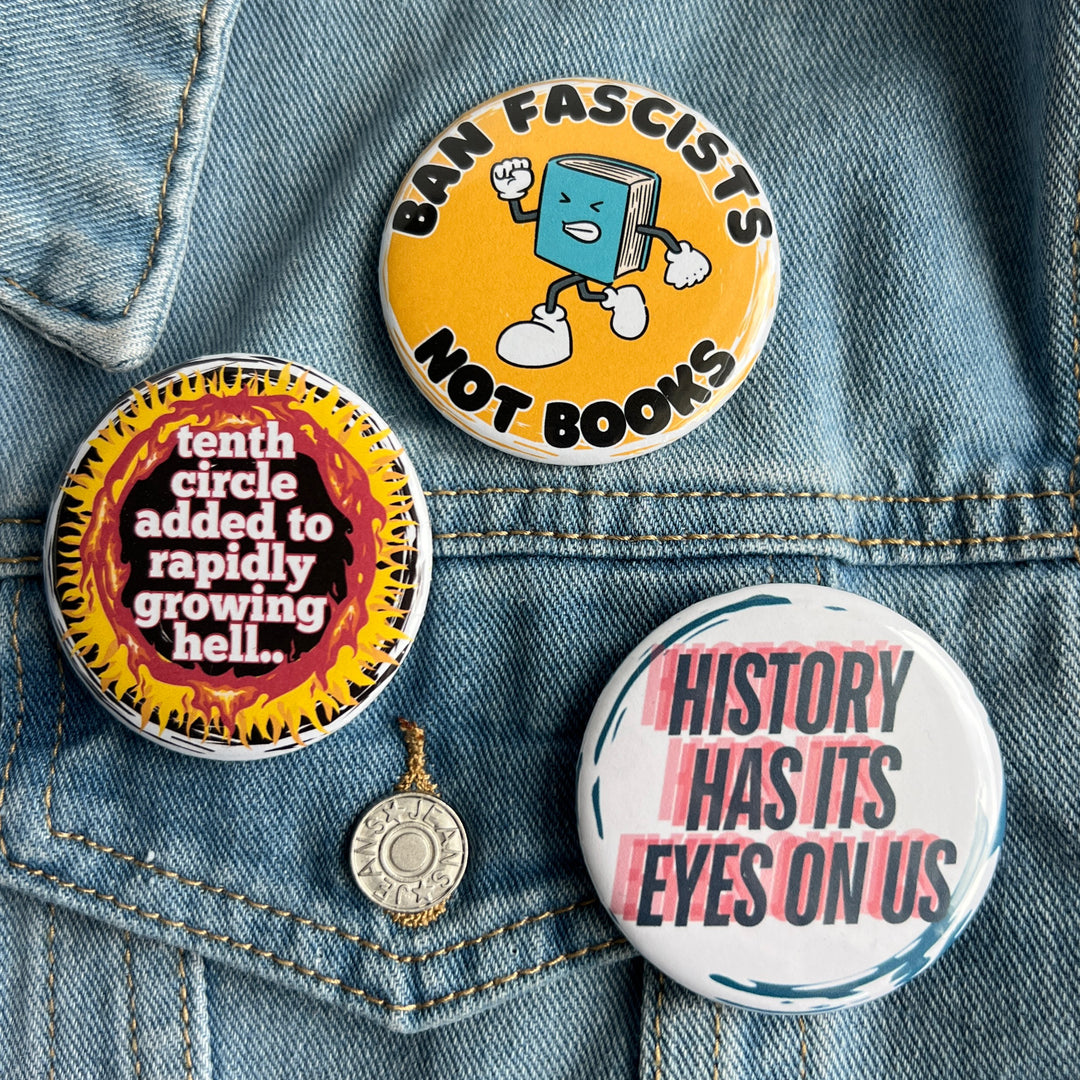 round pinback button with a black background, yellow and red flames surrounding the words, "tenth circle added to rapidly growing hell." A second button with a yellow background and a cartoon image of an angry book has the words, 'ban fascists not books.' A third button has a white background and the words, 'history has its eyes on us' in blue and red.  All pinned to a jean jacket for display.