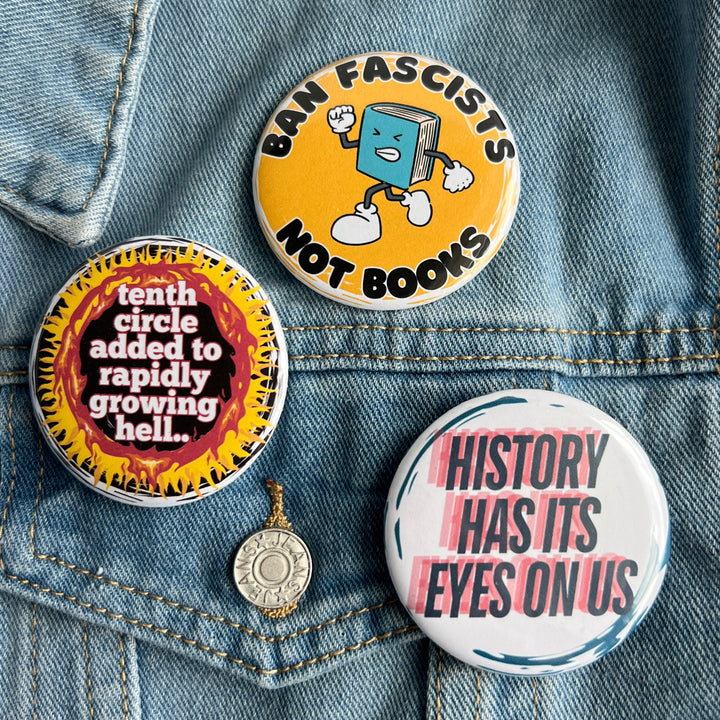 round pinback button with a black background, yellow and red flames surrounding the words, "tenth circle added to rapidly growing hell." A second button with a yellow background and a cartoon image of an angry book has the words, 'ban fascists not books.' A third button has a white background and the words, 'history has its eyes on us' in blue and red.  All pinned to a jean jacket for display.