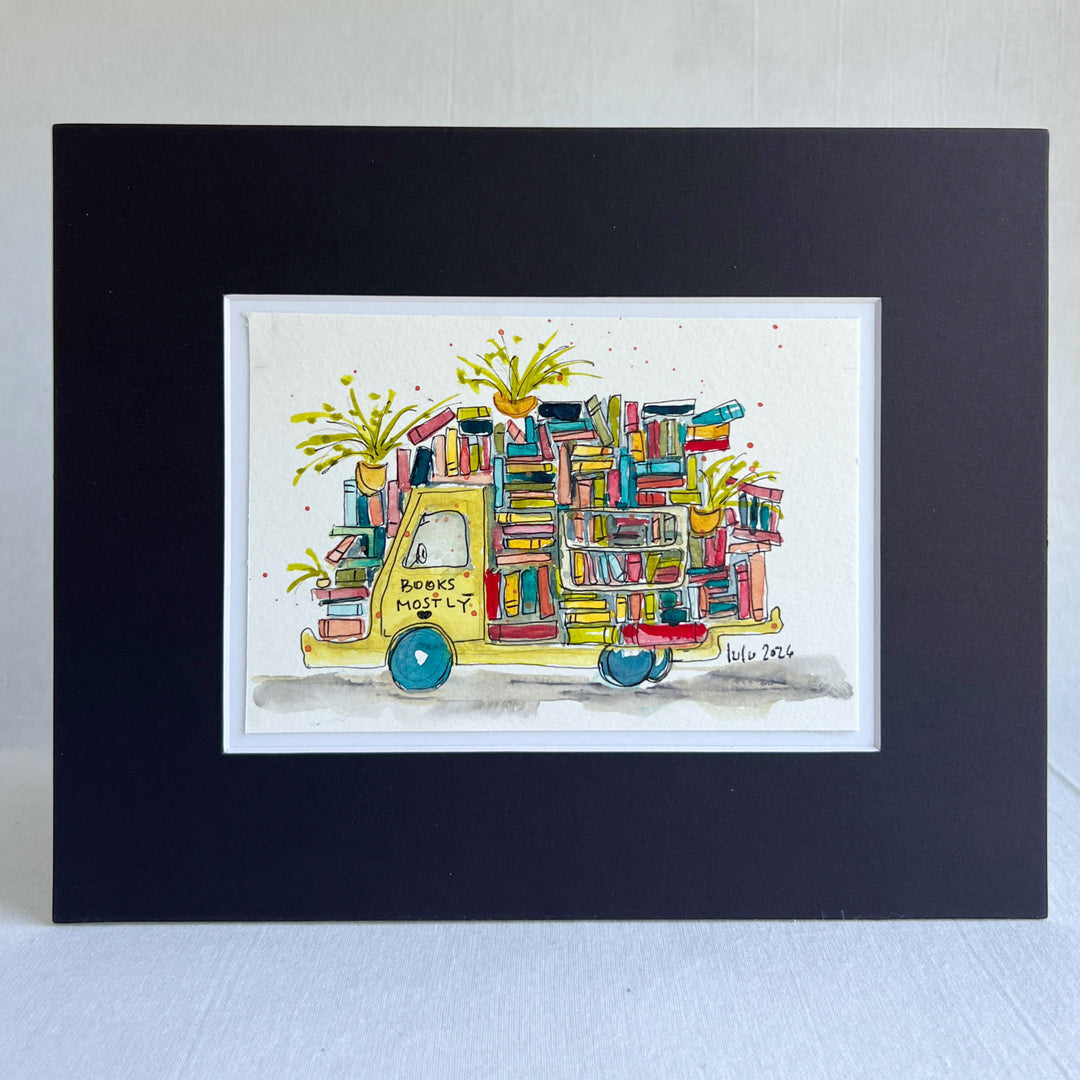 A framed watercolor art print featuring a yellow book truck filled with colorful books and potted plants, presented in a black frame with a white mat on a light textured background.
