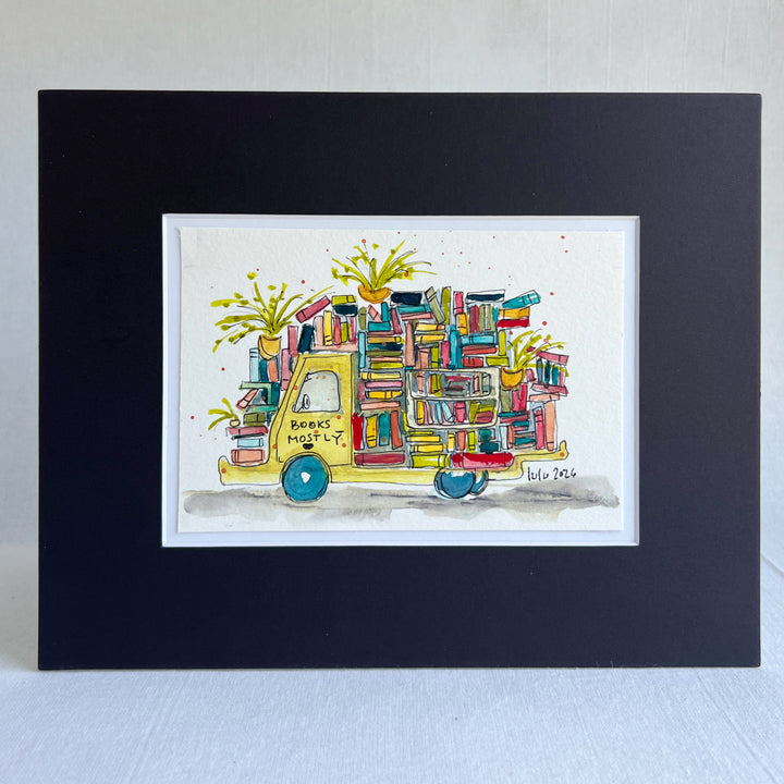 A framed watercolor art print featuring a yellow book truck filled with colorful books and potted plants, presented in a black frame with a white mat on a light textured background.