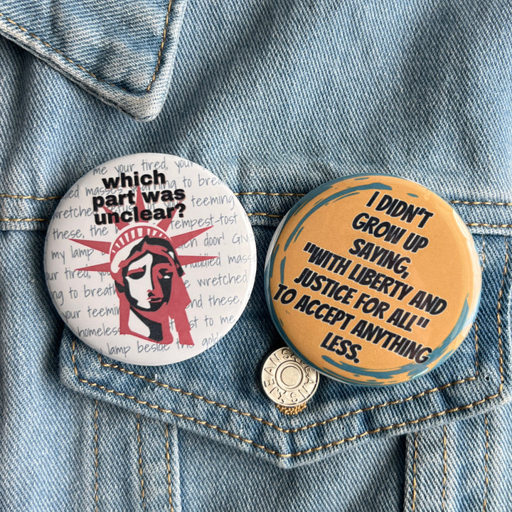 round pinback button with black lettering on a yellow background and the words, 'I didn't grow up saying "with liberty and justice for all" to accept anything less.' Pinned to a jean jacket for display. a second pinback button displays the statue of liberty in red and blue with the words, "which part was unclear?" referring to the background words displaying the engravings on the actual statue.