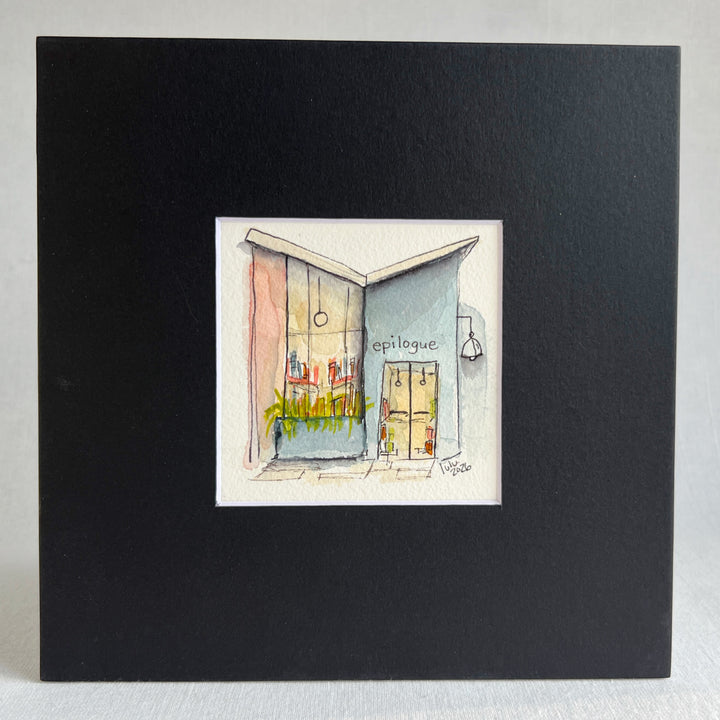 A matted watercolor painting of a cozy midcentury modern bookshop storefront with the store name  'EPILOGUE' above a window filled with books, a glass door, and a hanging lamp, shown on a white mat and white frame.