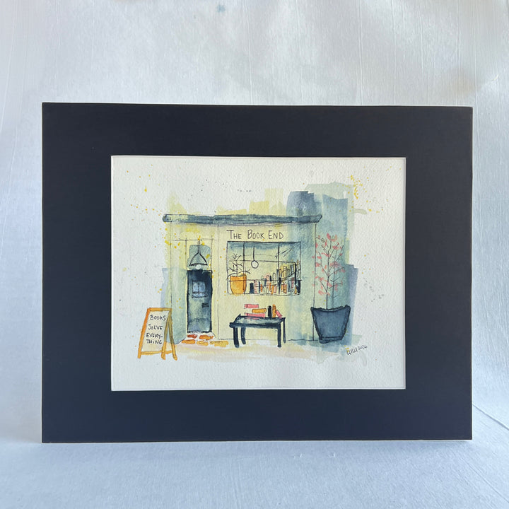 A watercolor painting of a mystery bookstore storefront labeled 'The Book End,' with a dark door, window display of books, a small table with books, potted plants, and a chalkboard sign reading 'Books Save Everything.' The artwork is painted on a white paper background with a dark navy border around it.