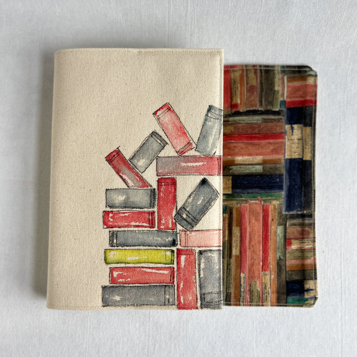 A rectangular canvas book cover featuring a watercolor illustration of stacked books in red, gray, and yellow on a beige background for the outside, and colorful book fabric for the inside.