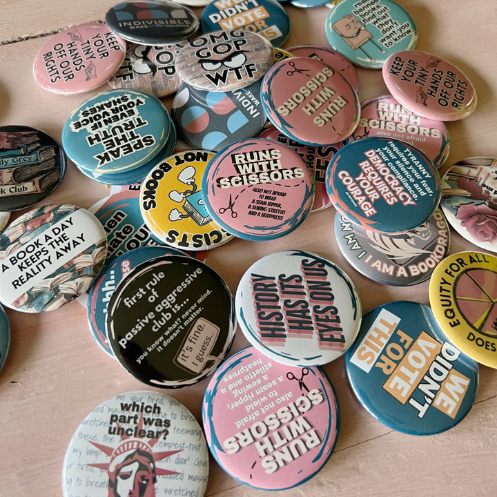 a photo of multiple pinback buttons in a variety of colors and quotes, scattered on a pink table for display.