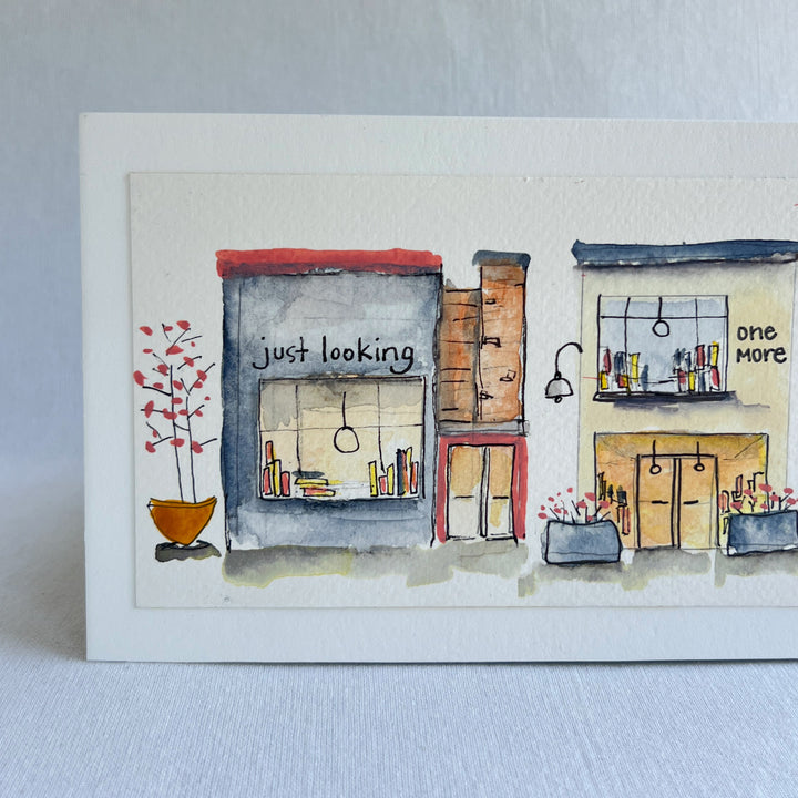 A watercolor painting of four colorful brick storefront bookstores, with the names, "Just Looking"  "One More" and "Last Chapter."  Displayed on top of a flower-painted book.