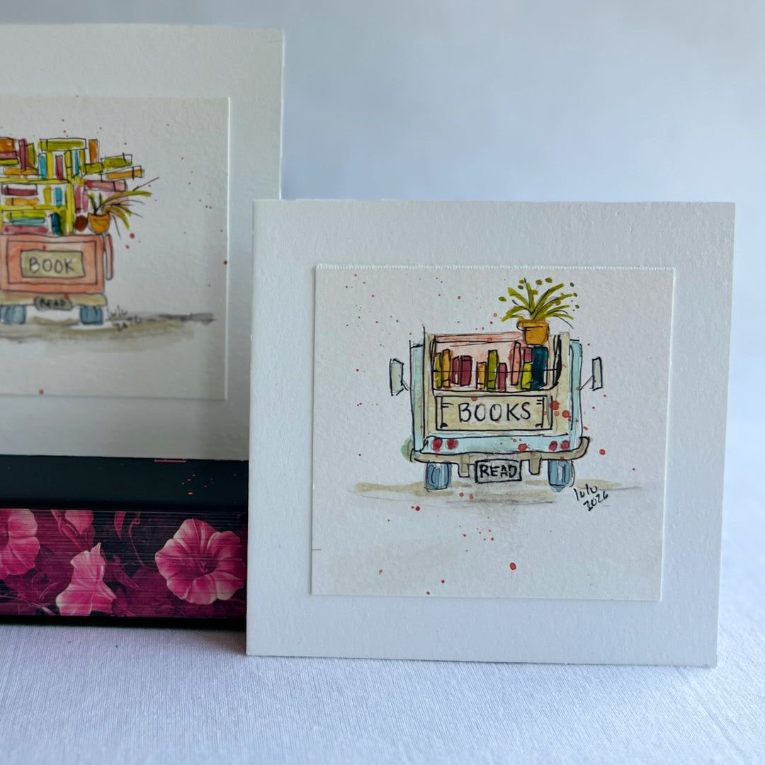 A square watercolor original art print featuring a hand-drawn and hand-painted illustration of a book cart with the word 'BOOKS' on the back and a small plant on top, displayed upright on stacked books.