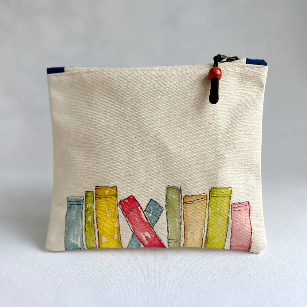 A canvas zippered pouch in a warm cream color, decorated with a watercolor illustration of colorful book spines along the bottom. The pouch has a black zipper with a red and black bead pull.