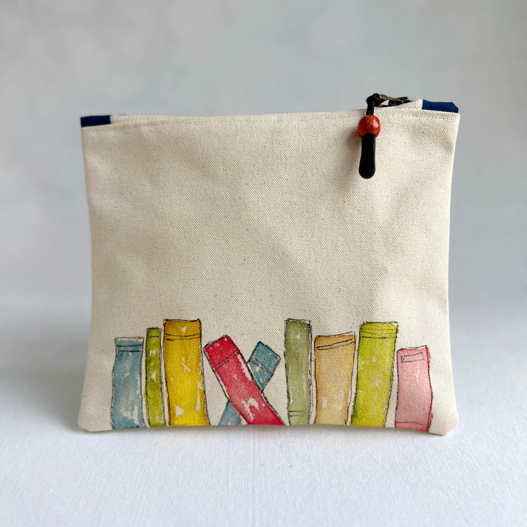 A canvas zippered pouch in a warm cream color, decorated with a watercolor illustration of colorful book spines along the bottom. The pouch has a black zipper with a red and black bead pull.