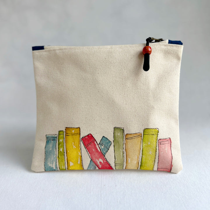 A canvas zippered pouch in a warm cream color, decorated with a watercolor illustration of colorful book spines along the bottom. The pouch has a black zipper with a red and black bead pull.