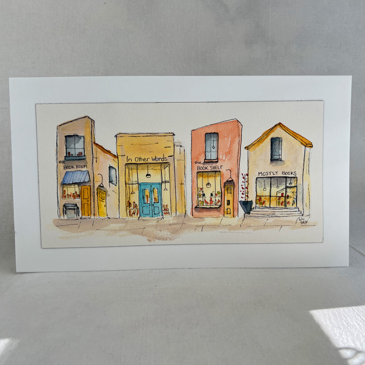 A watercolor art print depicting four stone bookstore buildings, presented with a wide white mat board against a neutral gray background.