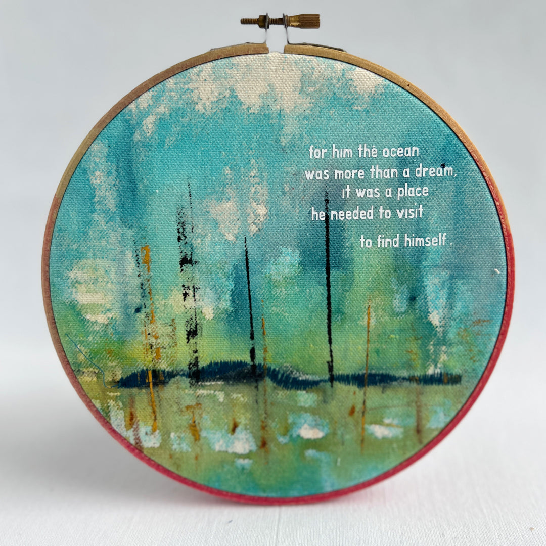 A large painted hoop art piece, with bright blues, citron green and touches of white. stitched with a deep blue shoreline and finished with the words, for him, the ocean was more than a dream. it was a place he needed to visit to find himself. 