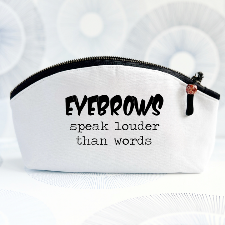 white canvas cosmetic bag with a curvy top, black zipper and the words, eyebrows speak louder than words.