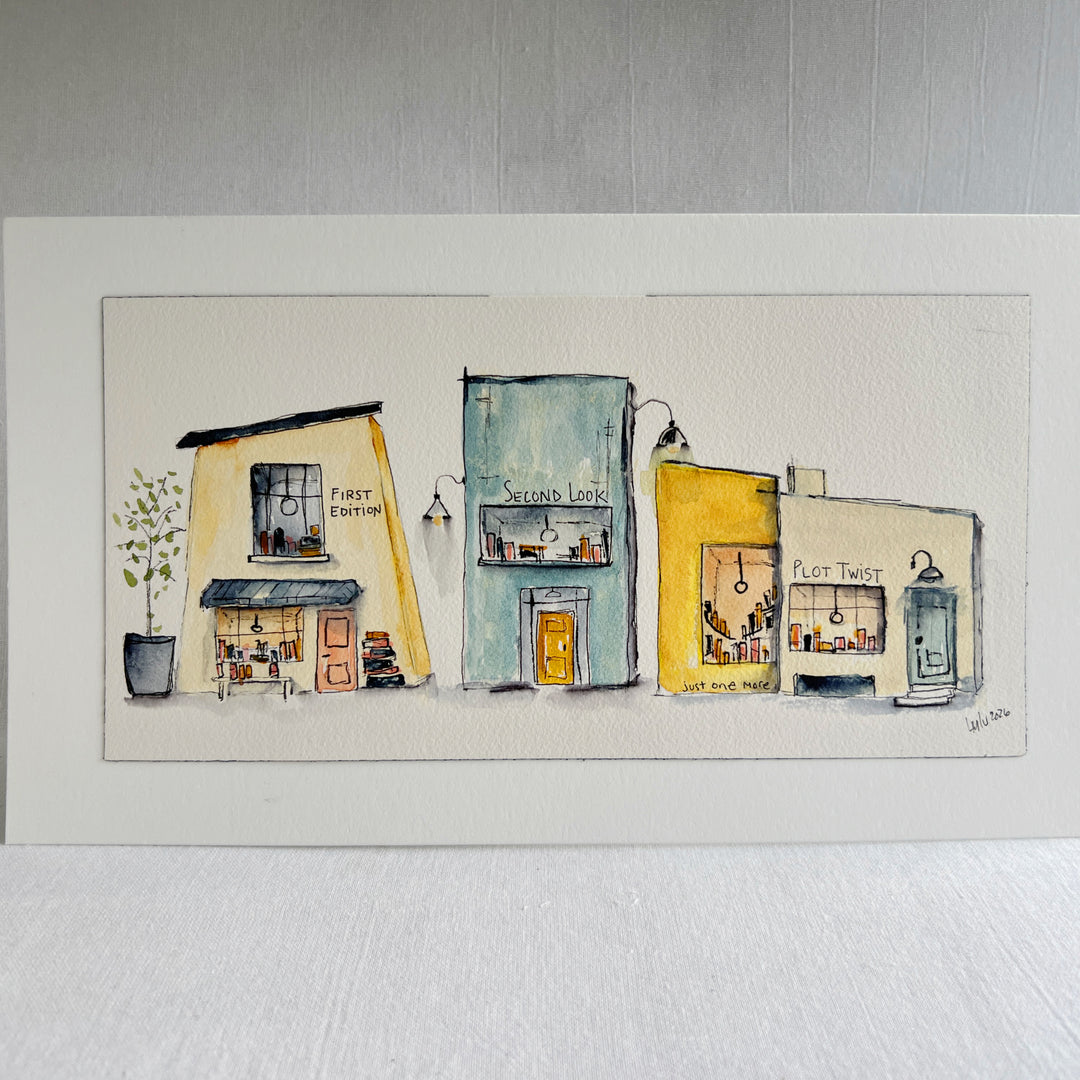 A horizontal watercolor painting of fthree whimsical storefront buildings in pastel yellow, blue, and cream tones, displayed on a white mat against a light gray textured wall.