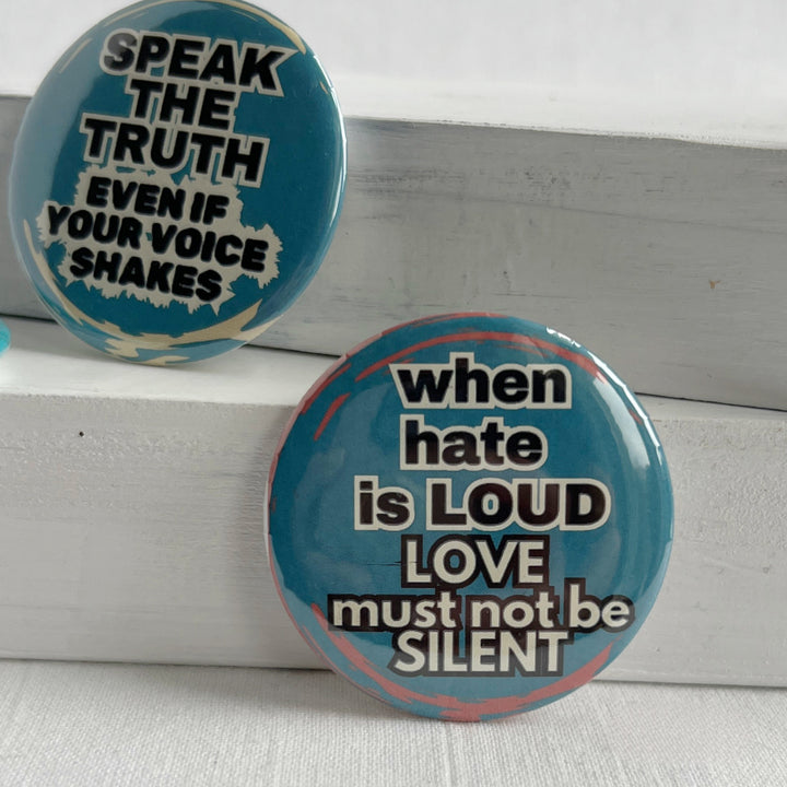 a round pinback button with a blue background and streaks of read, with the words, "when hate is LOUD, LOVE must not be SILENT."  Also pictured is a round pinback button with a blue background and the words, "Speak the Truth, even if your voice shakes."