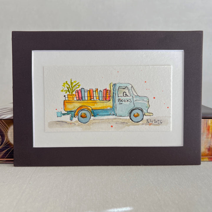 A matted watercolor art print featuring a light blue vintage truck carrying colorful books and a green plant, displayed in a black frame with a white mat on a tabletop.