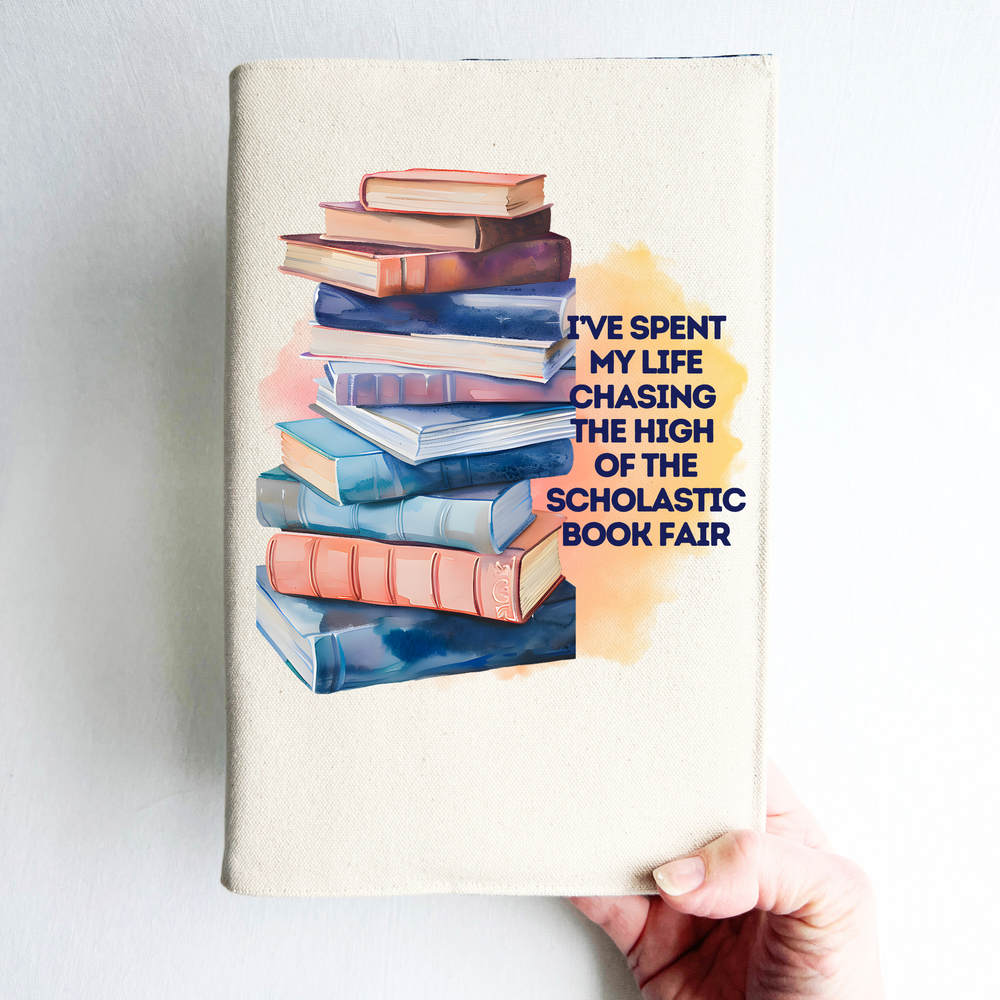Canvas, reusable book cover in linen, with the words,  I've spent my life chasing the high of the scholastic book fair' in navy lettering on a watercolor swoosh, next to a stack of books rendered in watercolor-painting style