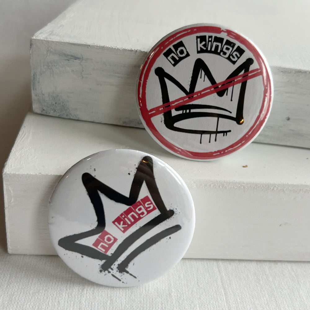a round, white pinback button with a black crown and a red circle with slash and the words, 'no kings.'  A second pinback button has a black crown and the words, 'no kings' in red.