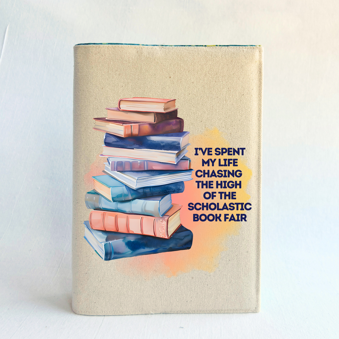 Canvas, reusable book cover in linen, with the words,  I've spent my life chasing the high of the scholastic book fair' in navy lettering on a watercolor swoosh, next to a stack of books rendered in watercolor-painting style