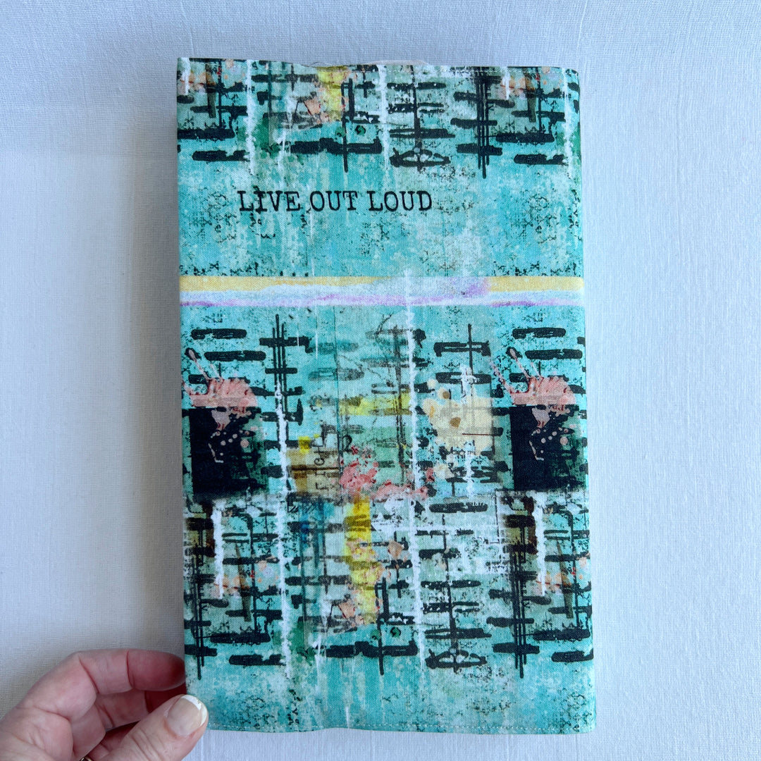 a book covered in a vibrant blue green fabric reusable book cover with the words, 'live out loud' printed on the top.
