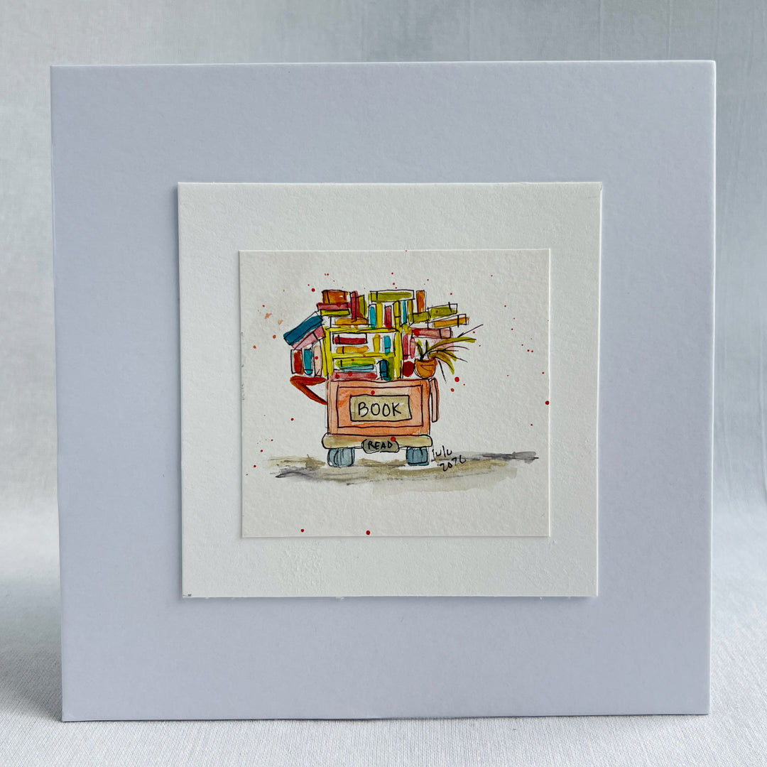 A square watercolor art print depicting a small book cart overfilled with colorful books and a potted plant, with the word “BOOK” written on the front.