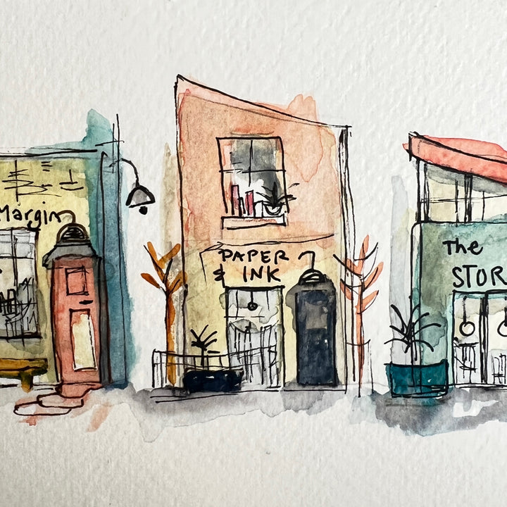 A horizontal watercolor art print depicting four whimsical bookstores with names including 'The Backlist', 'In the Margin, 'Paper & Ink', and 'The Story Shop'. The print is displayed on a textured light gray surface against a neutral gray background.