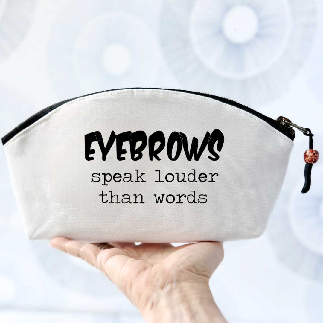 white canvas cosmetic bag with a curvy top, black zipper and the words, eyebrows speak louder than words.