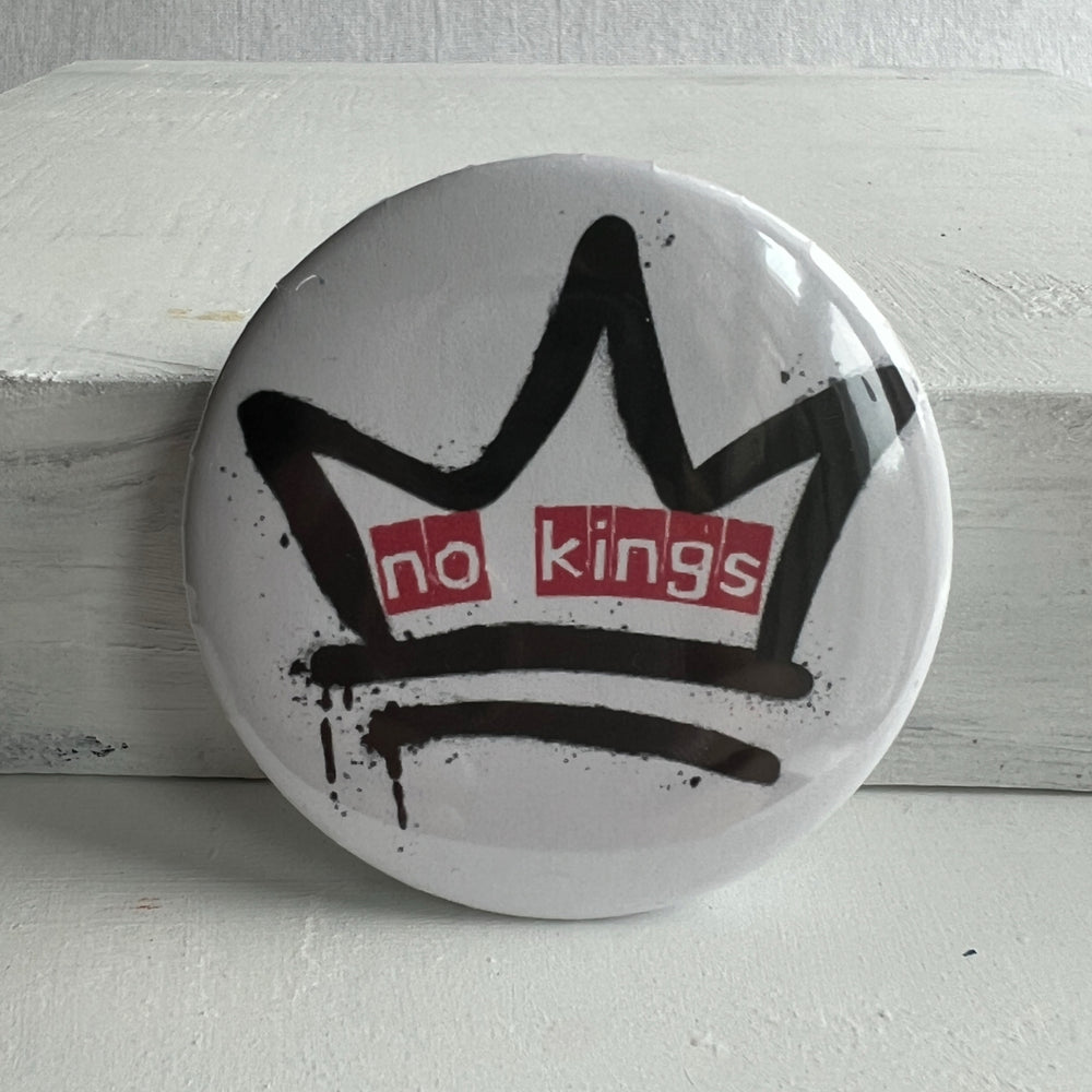 a round, white pinback button with a black crown and the words, 'no kings' in red.
