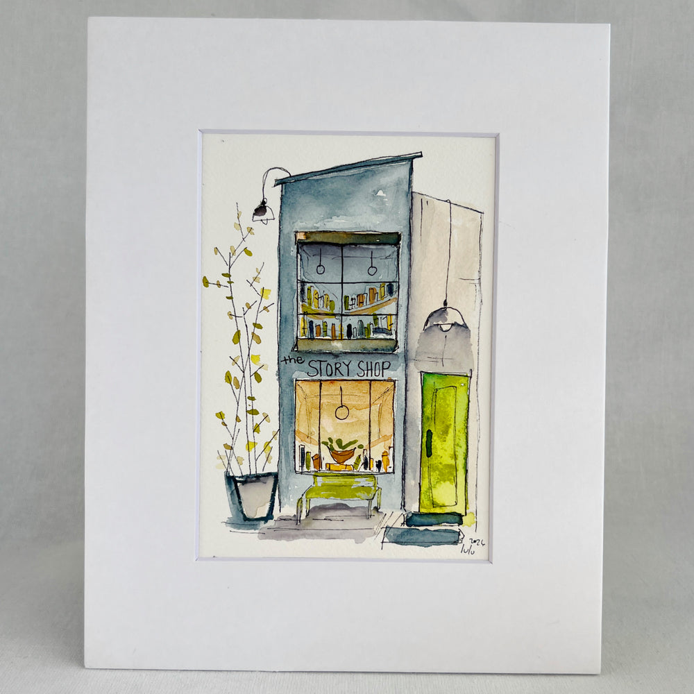 An original watercolor painting depicting a cozy, tall, narrow bookstore with the name "the Story Shop," with shelves, a green door, a bench, and a potted plant, presented in a white frame against a light gray background.