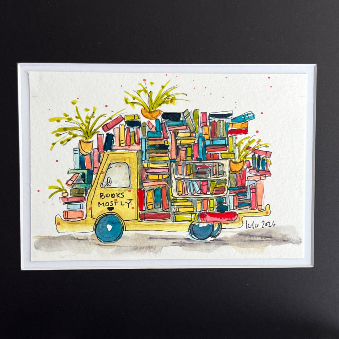 A framed watercolor illustration of a yellow truck carrying colorful books and potted plants, with the words 'BOOKS MOSTLY' on the side, shown against a dark background.