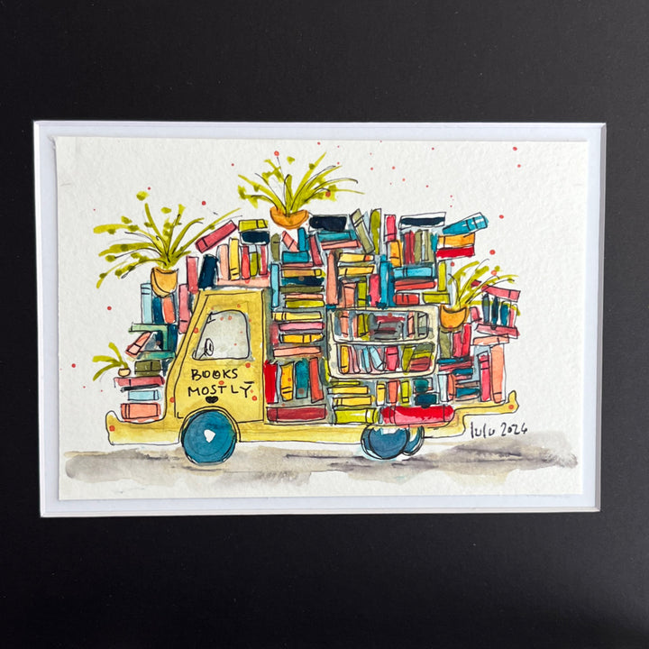 A framed watercolor illustration of a yellow truck carrying colorful books and potted plants, with the words 'BOOKS MOSTLY' on the side, shown against a dark background.