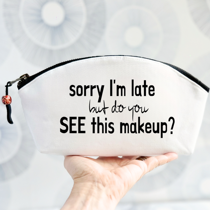 white canvas zip bag with black zipper, curved top and the words, sorry I'm late but do you see this makeup?