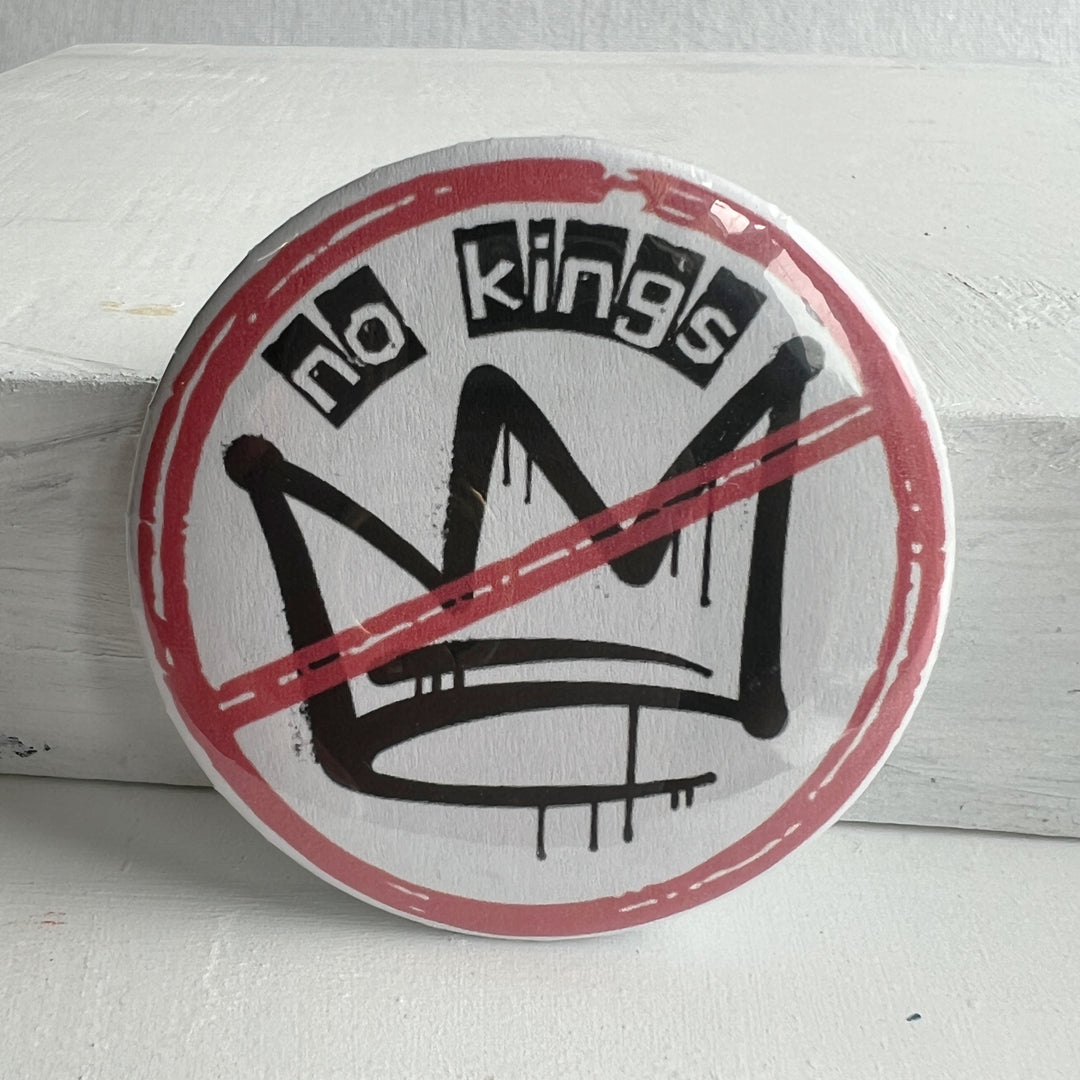a round, white pinback button with a black crown and a red circle with slash and the words, 'no kings.'
